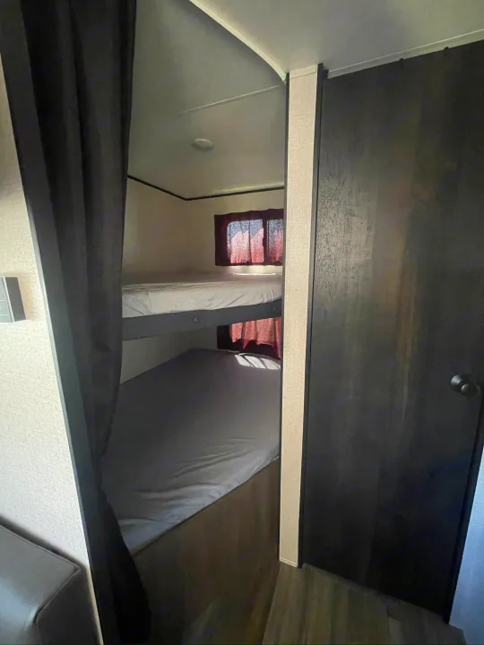 RV rental bunk bed area: stacked beds with light bedding, red-curtained windows, and a dark privacy curtain. Dark door on right.. Starcraft Autumn Ridge 2023