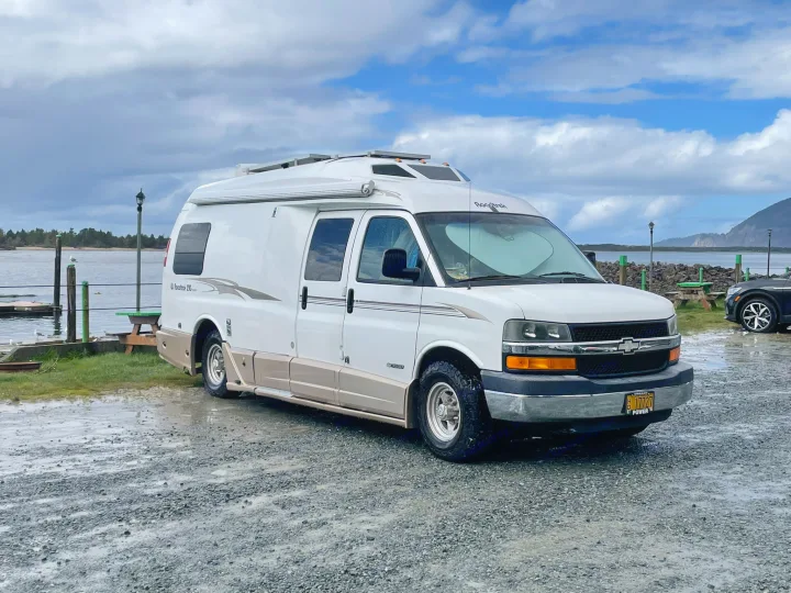 White Roadtrek RV rental parked on wet gravel by a bay, with solar panels visible on top, under a cloudy sky.. Roadtrek 210 - Popular 2005