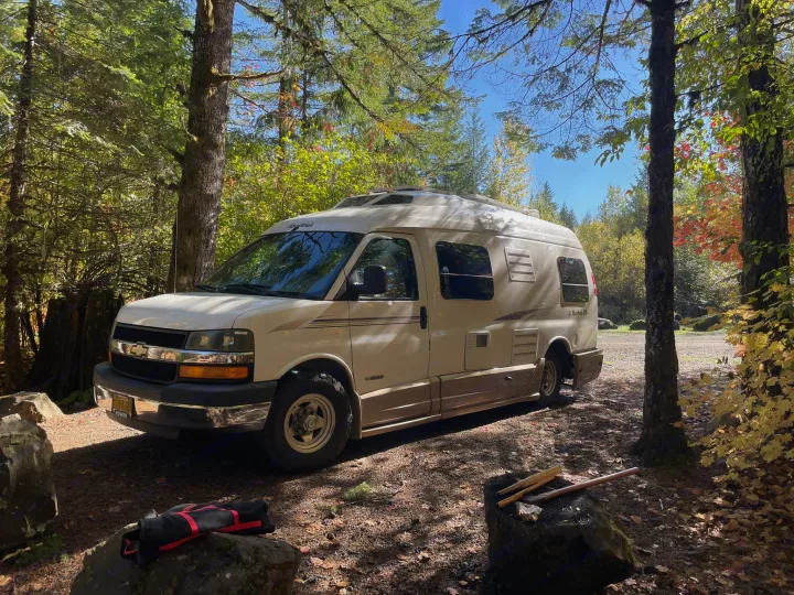 A white and tan RV rental is parked in a sunny, autumn-dappled forest campsite, ideal for nature exploration.. Roadtrek 210 - Popular 2005