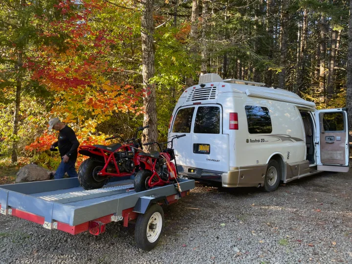 A white Roadtrek RV rental is parked with a trailer carrying mini bikes, surrounded by vibrant fall trees.. Roadtrek 210 - Popular 2005