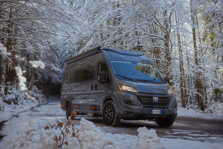 A grey RV rental, a camper van, is shown on a snow-covered forest road, flanked by snow-laden trees, ideal for winter adventures.. ELNAGH DUO XL 2025