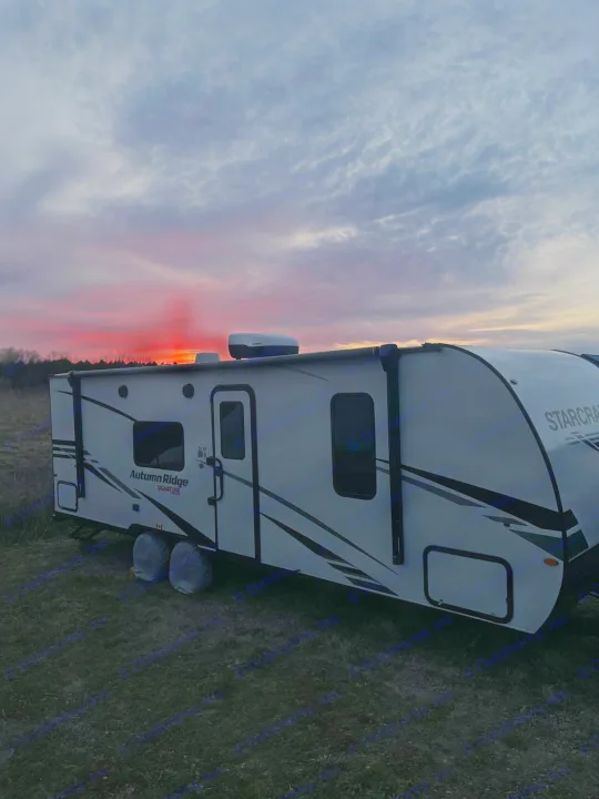 A beautiful red sunset illuminates a white Starcraft Autumn Ridge RV rental travel trailer in an open field.. Starcraft Autumn Ridge 2023
