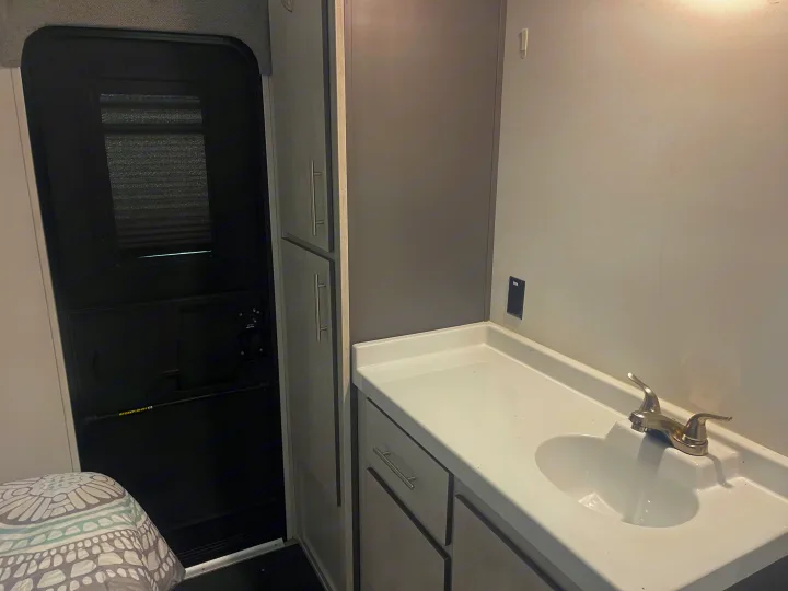 This RV rental shows a clean white vanity with a sink, silver faucet, light gray cabinets, and a bed by the dark entrance door.. Travel Lite Evoke 2020