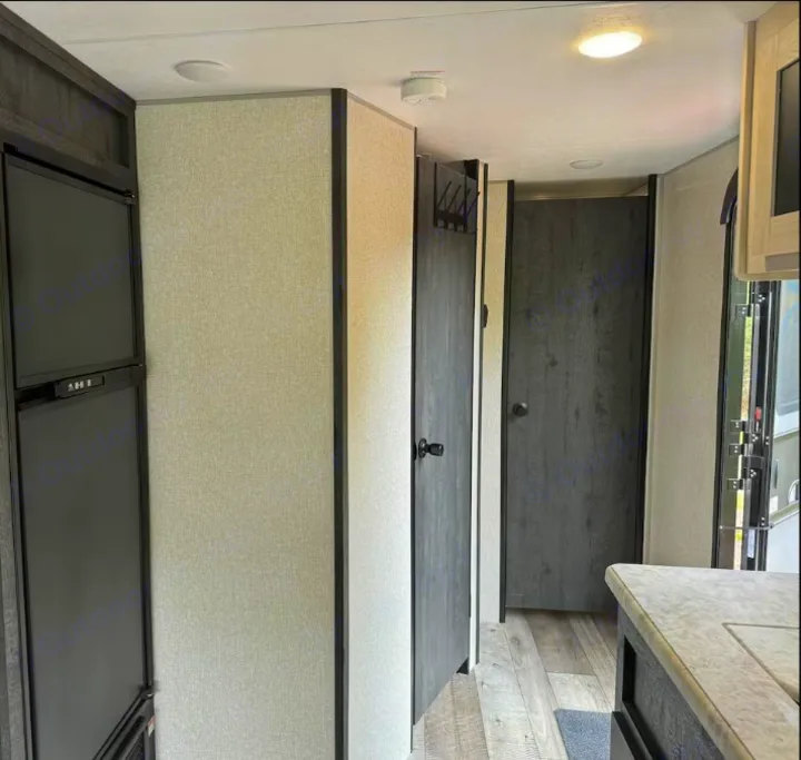 This RV rental interior shows a modern hallway with a dark fridge on the left, two dark wood doors, and a kitchen counter on the right.. Sportsmen by KZ 250THLE 2022