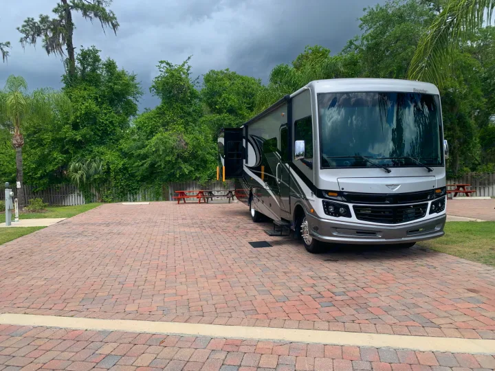 A large grey and black RV rental is parked on a brick paver site, surrounded by green trees under a cloudy sky at a campsite.. Fleetwood Bounder 33C 2021