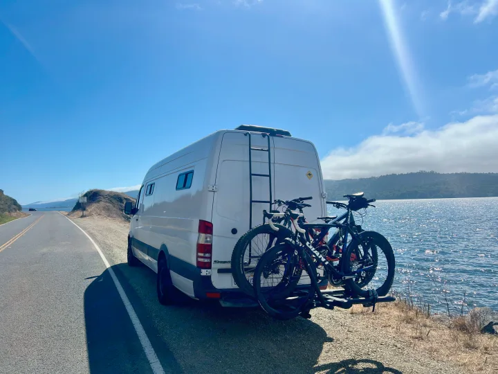 A white Sprinter van, an RV rental, with bikes on the back, parked on a road next to sparkling water on a sunny day.. Dodge Sprinter 3500 2007