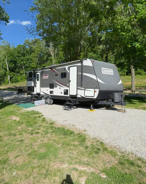 Grey and white Coleman RV rental parked on a gravel site, surrounded by green trees under a blue sky, ready for adventure.. Dutchmen Coleman Lantern 2019