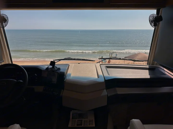 RV rental interior: dashboard & steering wheel. View of ocean, sandy beach, people in water & on shore, beach umbrella.. Fleetwood Bounder 33C 2021