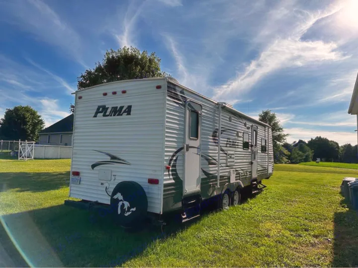 White Puma travel trailer RV rental, seen from the rear-side. Parked on green grass under a blue sky, ready for your next trip.. Forest River Palomino Puma 2013