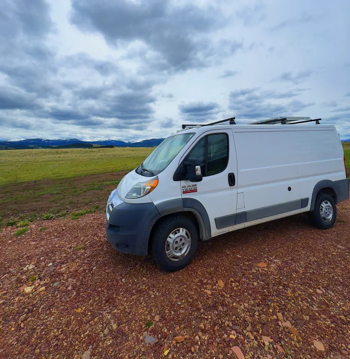 White Ram ProMaster camper van (RV rental) on a gravel road, backed by snowy mountains under cloudy skies. Explore the outdoors!. RAM Promaster 2017