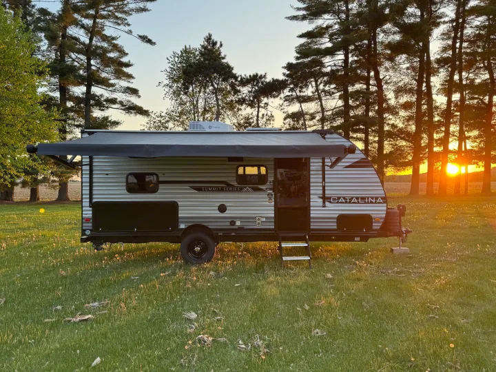 This RV rental travel trailer is parked in a grassy field with a warm sunset behind tall trees, ideal for a camping adventure.. Forest River 184BHS 2025