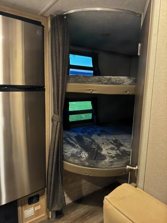 Compact RV rental interior: stainless steel fridge, two bunk beds with windows and curtains.. Forest River 184BHS 2025