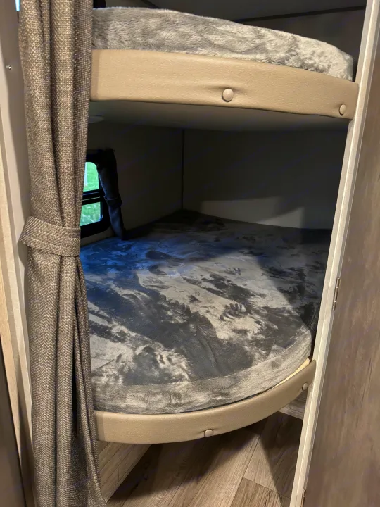 Inside an **RV rental**: view of cozy bunk beds with soft blankets, privacy curtain, and window, providing a compact sleeping area.. Forest River 184BHS 2025