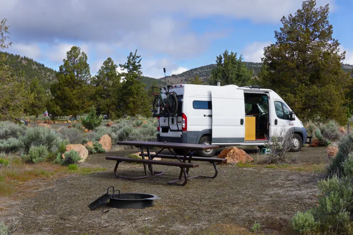 An rv rental van is parked at a scenic campsite with a picnic table and fire pit, surrounded by trees and mountains.. Ram promaster 2500 2017
