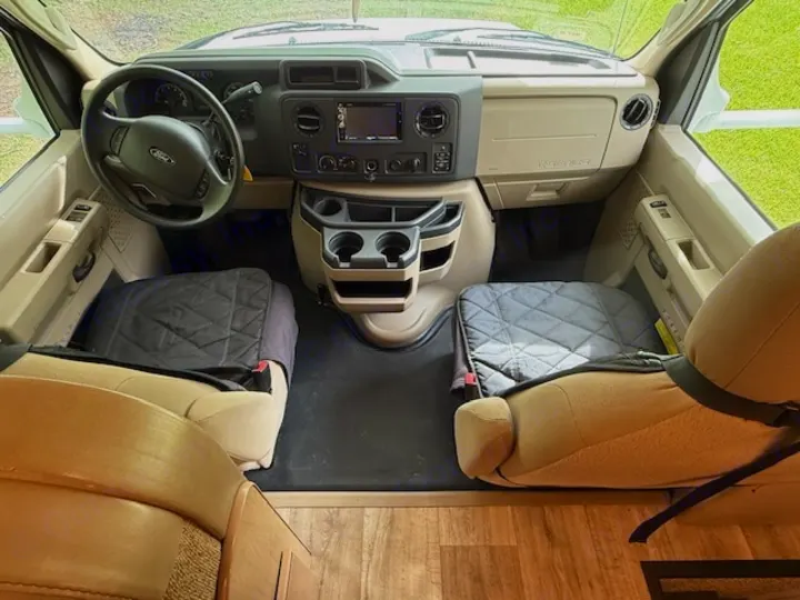 RV rental driver's cabin interior. Beige seats with quilted covers, Ford steering wheel, dashboard, and central console visible.. Thor Freedom Elite 2017