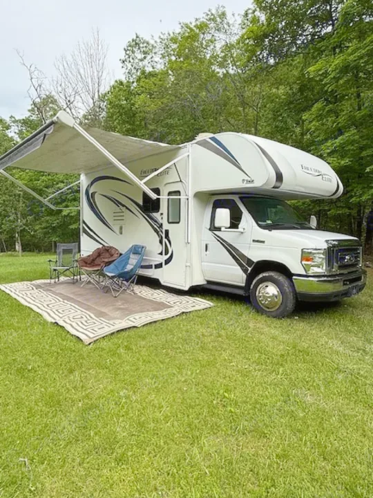 White Class C RV rental on grass with an extended awning, outdoor rug, and chairs, set up for camping.. Thor Freedom Elite 2017