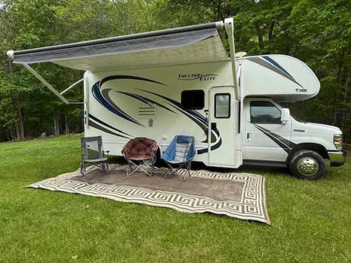 White Class C rv rental with extended awning, outdoor rug, and chairs on a grassy campsite.. Thor Freedom Elite 2017