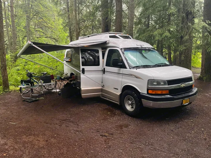 Roadtrek RV rental parked at a forest campsite with awning, chairs, and bike setup.. Roadtrek 210 - Popular 2005