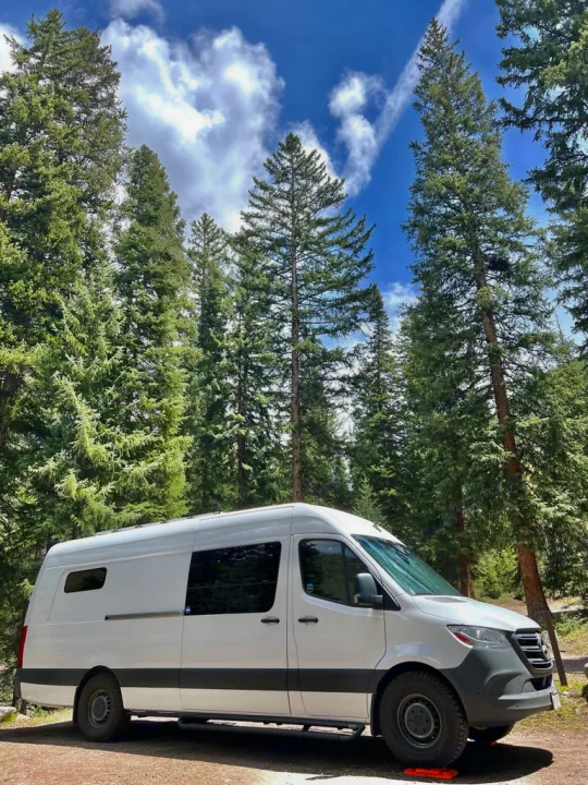 A white Sprinter camper van is parked among tall pine trees under a blue sky. Perfect RV rental for nature adventures.. Mercedes-Benz Sprinter 2020