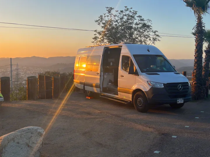 White Sprinter RV rental parked at a scenic sunset. Side door open, sunlight reflecting off the van, mountains in the distance.. Mercedes-Benz Sprinter 2020