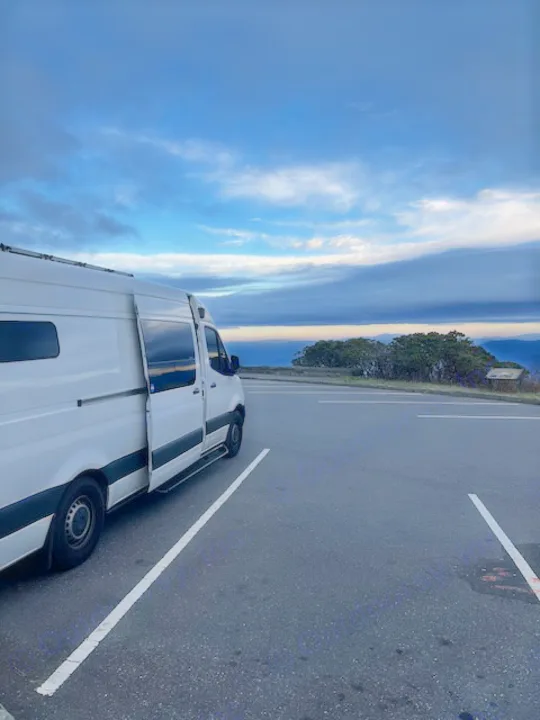 An RV rental, a white camper van, is parked with its side door open at a scenic mountain overlook under a cloudy sky.. Mercedes-Benz Sprinter 2020