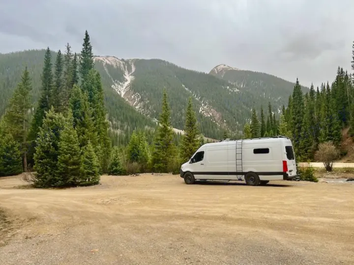 White rv rental camper van parked on dirt amidst a lush evergreen forest & mountains under an overcast sky.. Mercedes-Benz Sprinter 2020
