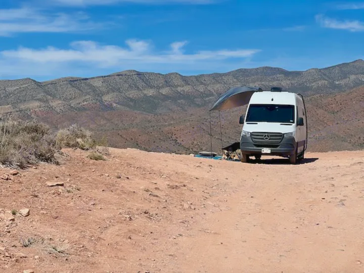 Explore the desert in comfort with an RV rental. A white camper van with an awning faces arid mountains under a blue sky.. Mercedes-Benz Sprinter 2020