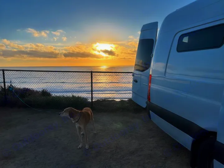 Scenic RV rental view: White camper van by ocean at sunset with dog on leash. Perfect for van life adventures.. Mercedes-Benz Sprinter 2020
