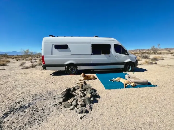 This rv rental van makes desert camping easy! A white camper van, fire pit, and 3 dogs relax on a blue mat in the sand.. Mercedes-Benz Sprinter 2020
