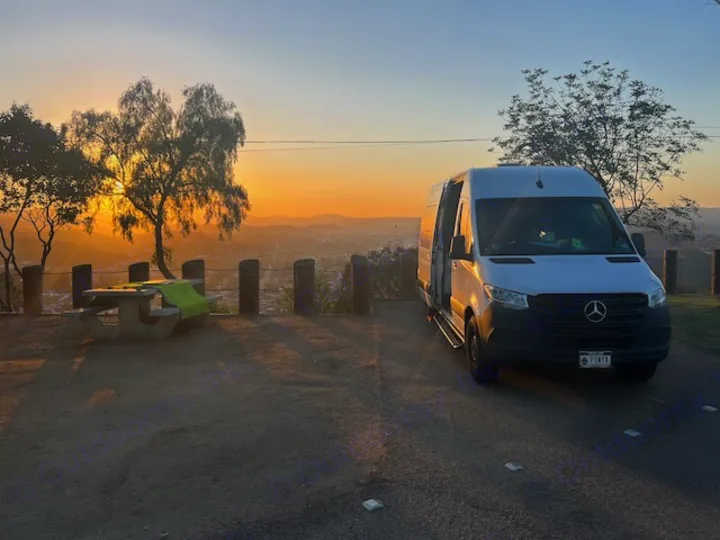 Golden hour illuminates an rv rental parked at a scenic overlook, offering stunning city views alongside a picnic table for travelers.. Mercedes-Benz Sprinter 2020