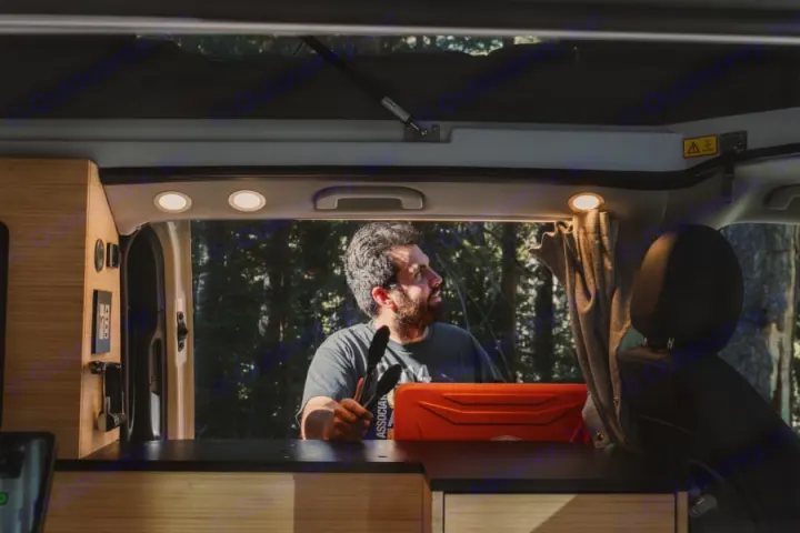 Inside an RV rental camper van, a man with tongs and an orange stove looks out at a forest, ready for outdoor cooking.. Mercedes-Benz metris 2022