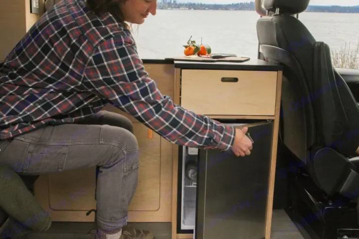 Enjoying the convenience of an RV rental: A person opens a compact fridge in a cozy camper van with a scenic lake view.. Mercedes-Benz metris 2022