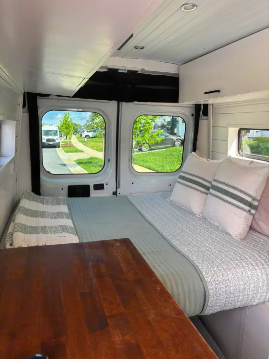 Cozy RV rental interior: bed with pillows, wooden table, and street views through large windows, perfect for travel.. Ram Ram ProMaster 2500 2019