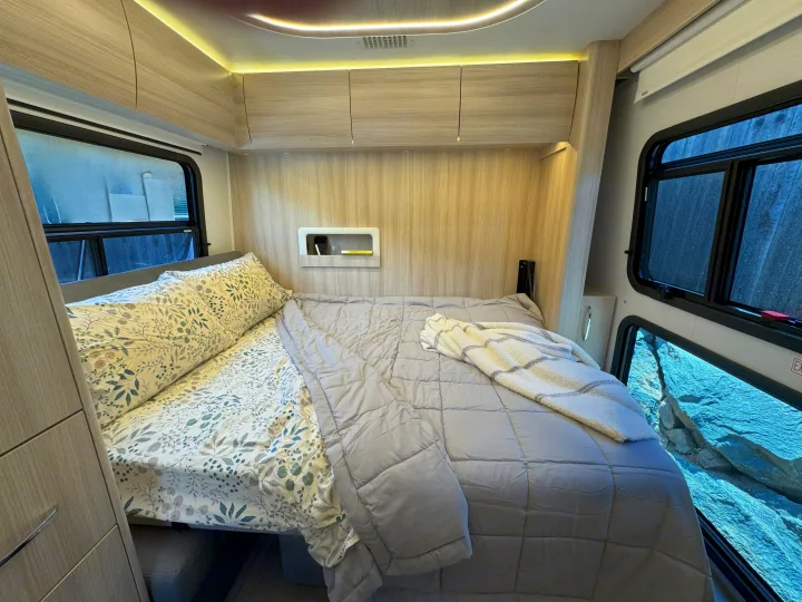 Cozy RV rental bedroom featuring a bed with patterned sheets, light wood decor, and windows offering scenic views.. Leisure Travel Vans Unity 2022
