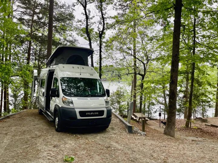 Enjoy your rv rental in nature! White camper van parked at a lakefront campsite with pop-top open, surrounded by trees.. Dodge Winnebago 2021