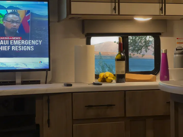 RV rental interior: TV shows "Maui Emergency Chief Resigns" news. Counter has wine, bananas, and a beautiful lake view outside the window.. Grand Design Imagine 2023