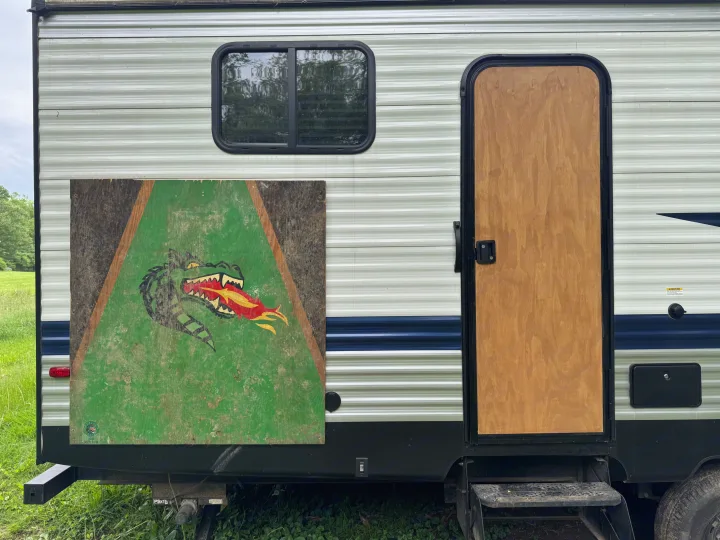 An RV rental's side with a fun dragon-themed cornhole board, entry door, and window. Perfect for outdoor adventures.. Keystone RV Springdale 2023