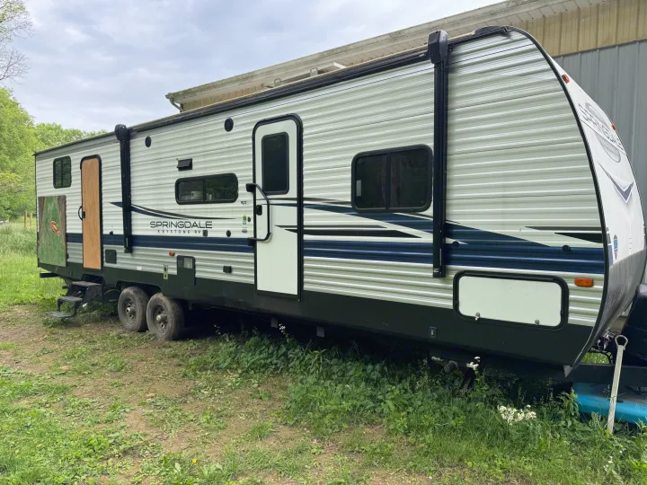 White and blue Springdale Keystone travel trailer, suitable for rv rental, parked on grass next to a building.. Keystone RV Springdale 2023