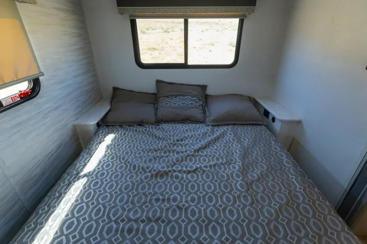 Inside an RV rental: a neatly made bed with grey geometric bedding, pillows, and a window overlooking an arid landscape.. Gulf Stream Conquest 2024