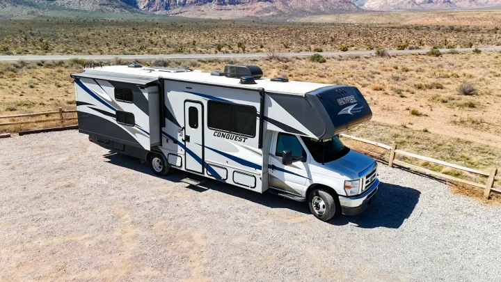 A white Class C rv rental with blue accents, parked on a gravel lot in a sunny desert scene with distant mountains.. Gulf Stream Conquest 2024