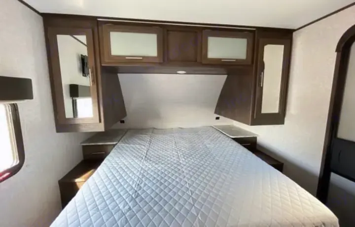 Inside an RV rental, a well-lit bedroom features a quilted bed, overhead cabinets, and side storage for comfortable travel.. Dutchmen Coleman Light 2020