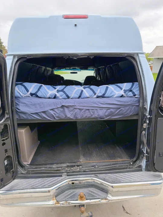 High-top van rv rental: converted interior with elevated bed & storage, seen through open rear doors. Tow hitch visible.. Ford E350 Super Duty 1-ton Camper Van 2006