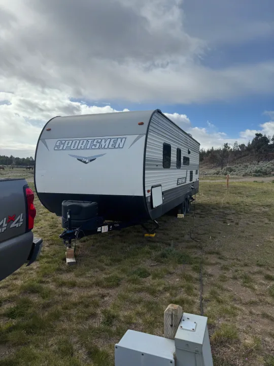 A white and grey Sportsmen travel trailer RV rental, connected to a truck, is parked in a grassy field with cloudy skies and distant horses.. KZ Sportsmen SE 260BHSE 2022