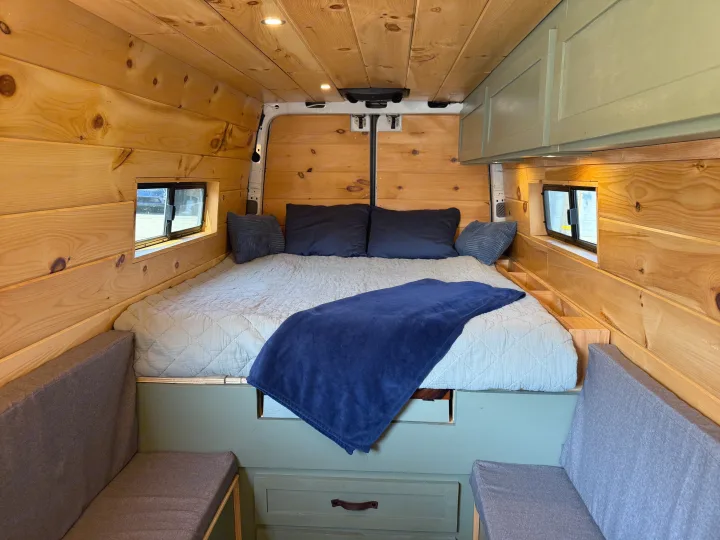 Cozy RV rental interior with a comfortable bed, warm wooden walls, and sage green cabinetry for a perfect camper van experience.. Dodge Sprinter 3500 2007