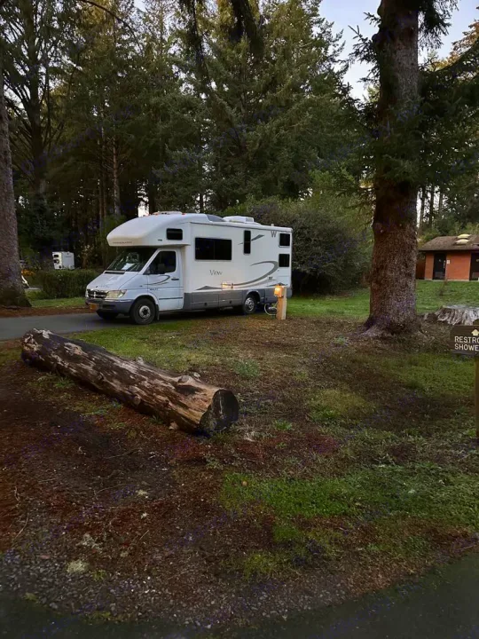 White RV rental parked at a quiet forest campsite among tall trees. A large log lies in the foreground, with a building behind.. Winnebago View 2006