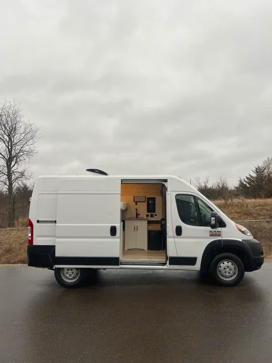 White camper van conversion with open side door revealing a custom kitchen and bed. Ideal RV rental for mobile adventures.. Ram Promaster 1500 2020