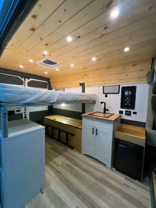 A custom RV rental interior featuring an elevated bed, compact kitchenette with sink, and ceiling lights.. Ram Promaster 1500 2020