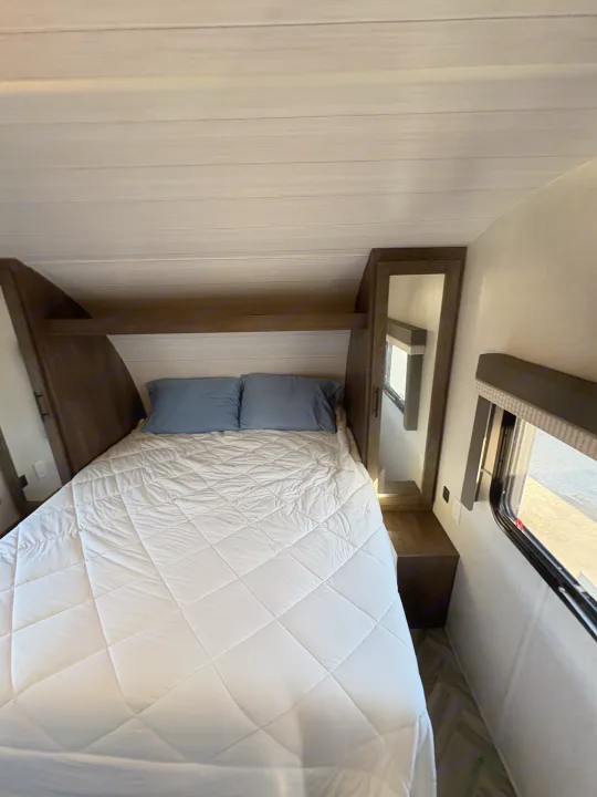 Cozy RV rental bedroom featuring a bed with blue pillows, white quilted comforter, wood-paneled ceiling, and a bright window.. Forest River Wildwood X-Lite 2022
