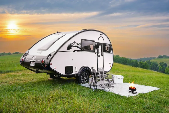 Enjoy nature with this teardrop RV rental, featuring a campfire setup under a beautiful sunset in a grassy field.. nuCamp T@B 2025