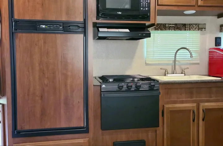 RV rental kitchen: a compact setup featuring wood-paneled fridge, microwave, gas range, and sink by a window.. Jayco Jay Flight SLX 2016